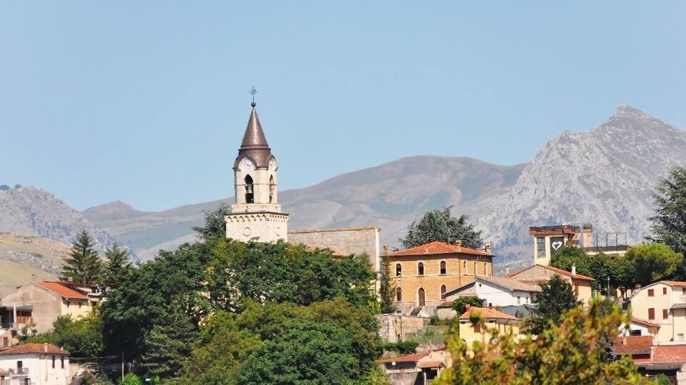 A town with a prominent church tower, surrounded by residential buildings and mountains in the background.