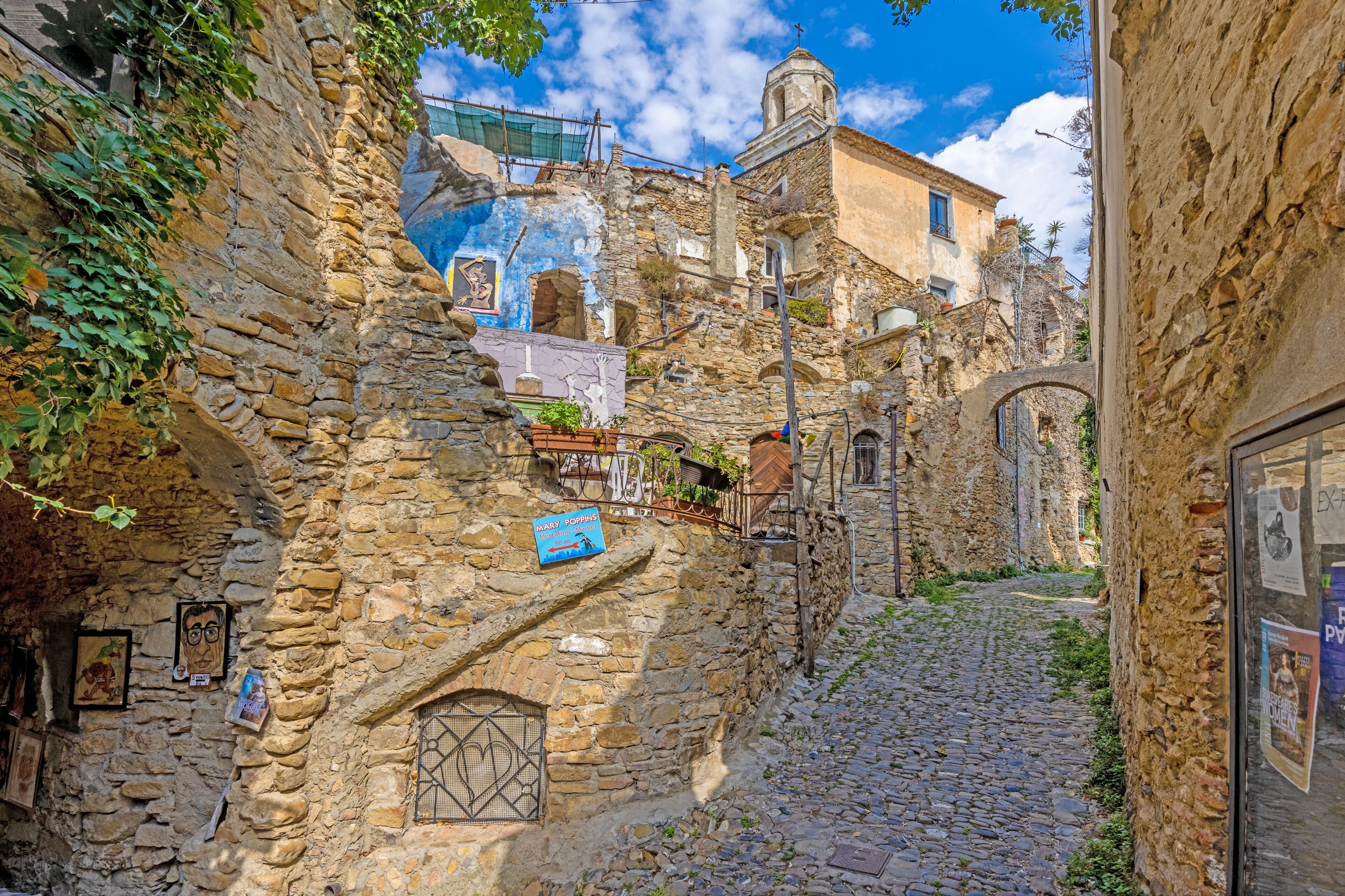A narrow cobblestone street flanked by stone buildings with signs and posters.