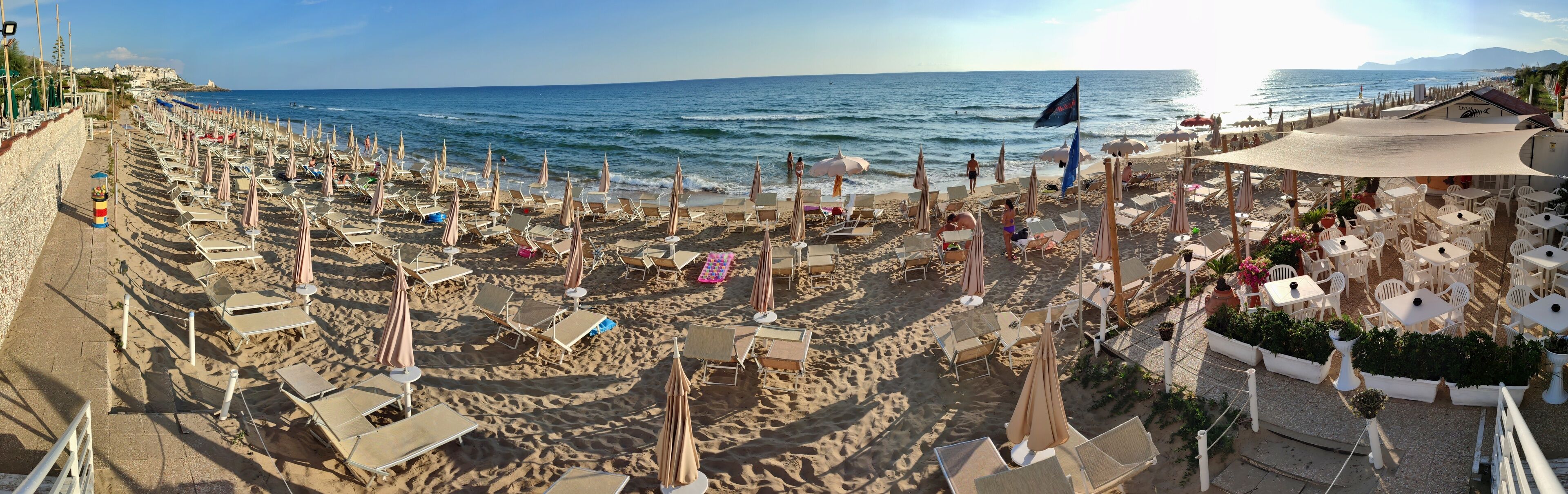 A beach with rows of sun loungers and umbrellas, a café area with outdoor seating, and the ocean in the background.