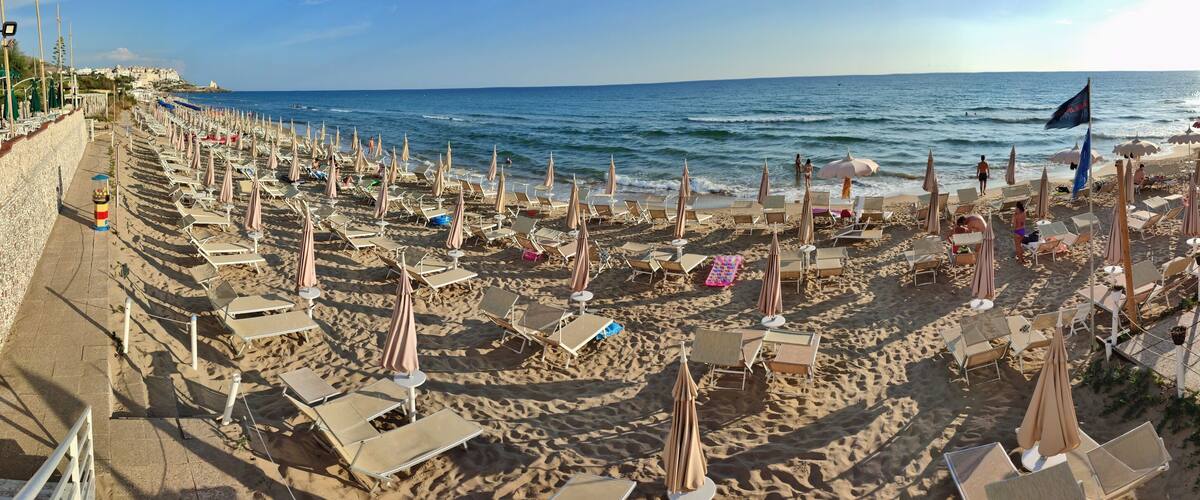 A beach with rows of sun loungers and umbrellas, a café area with outdoor seating, and the ocean in the background.