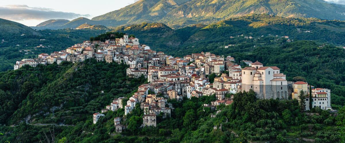 A hillside town with buildings nestled among lush green hills and mountains in the background.