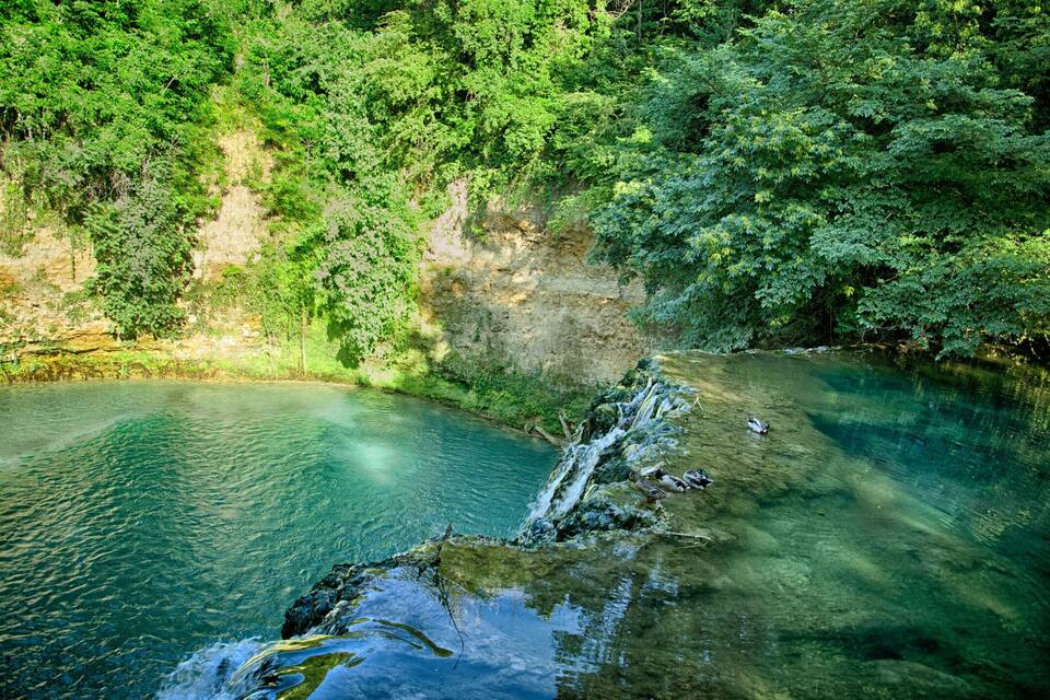 Een heldere, turquoise rivier die door een bebost gebied stroomt, met een rotsachtige klif en een waterval.