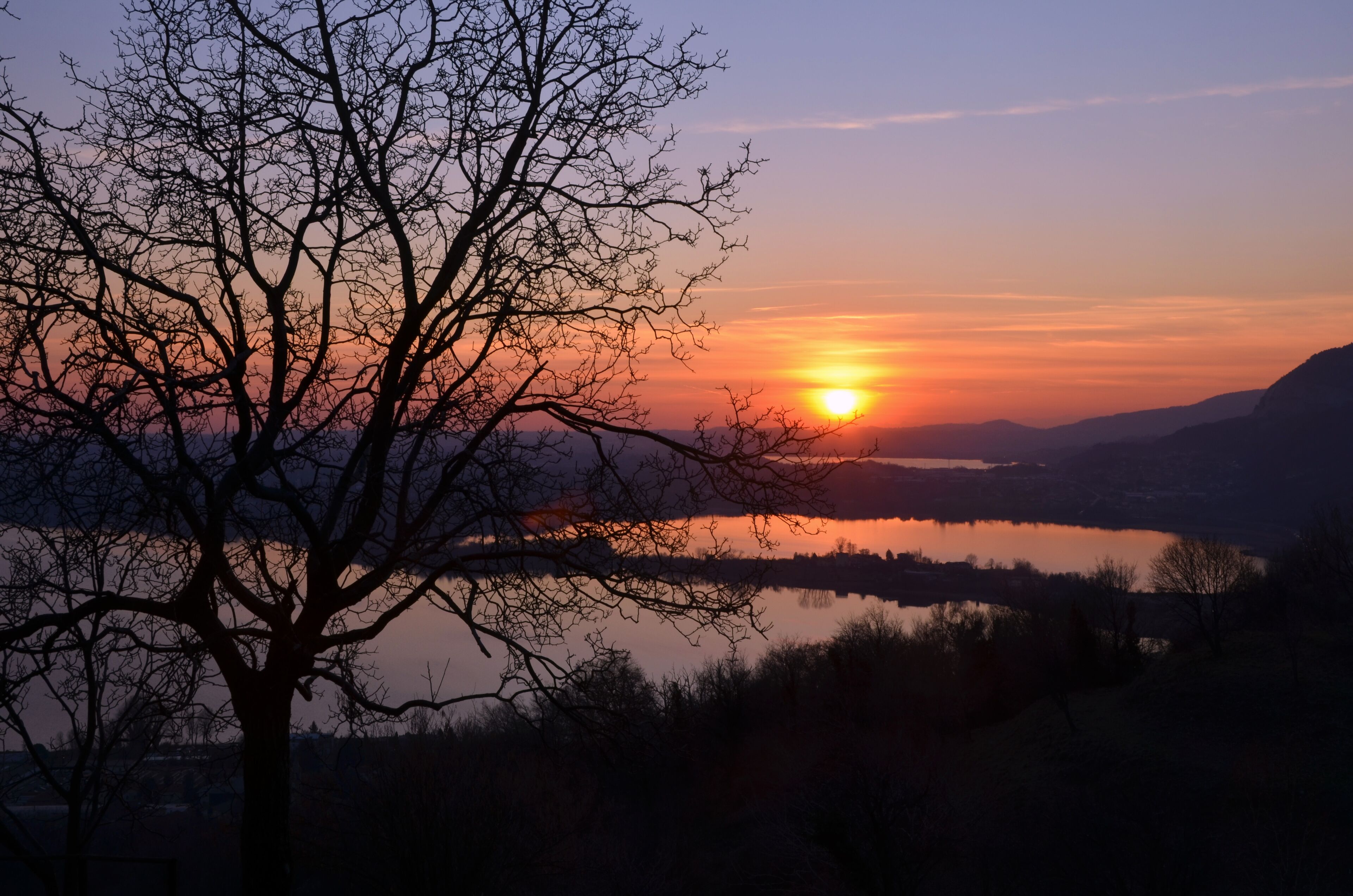A sunset over a calm lake with silhouetted trees and distant hills.