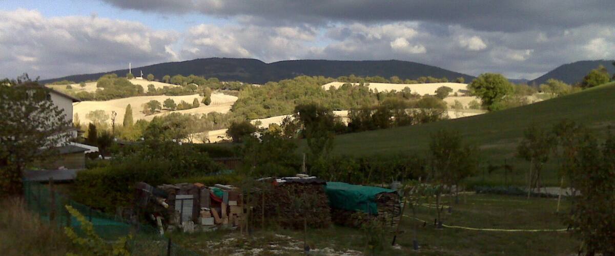 A rural landscape with rolling hills, a farmhouse, and a cloudy sky.
