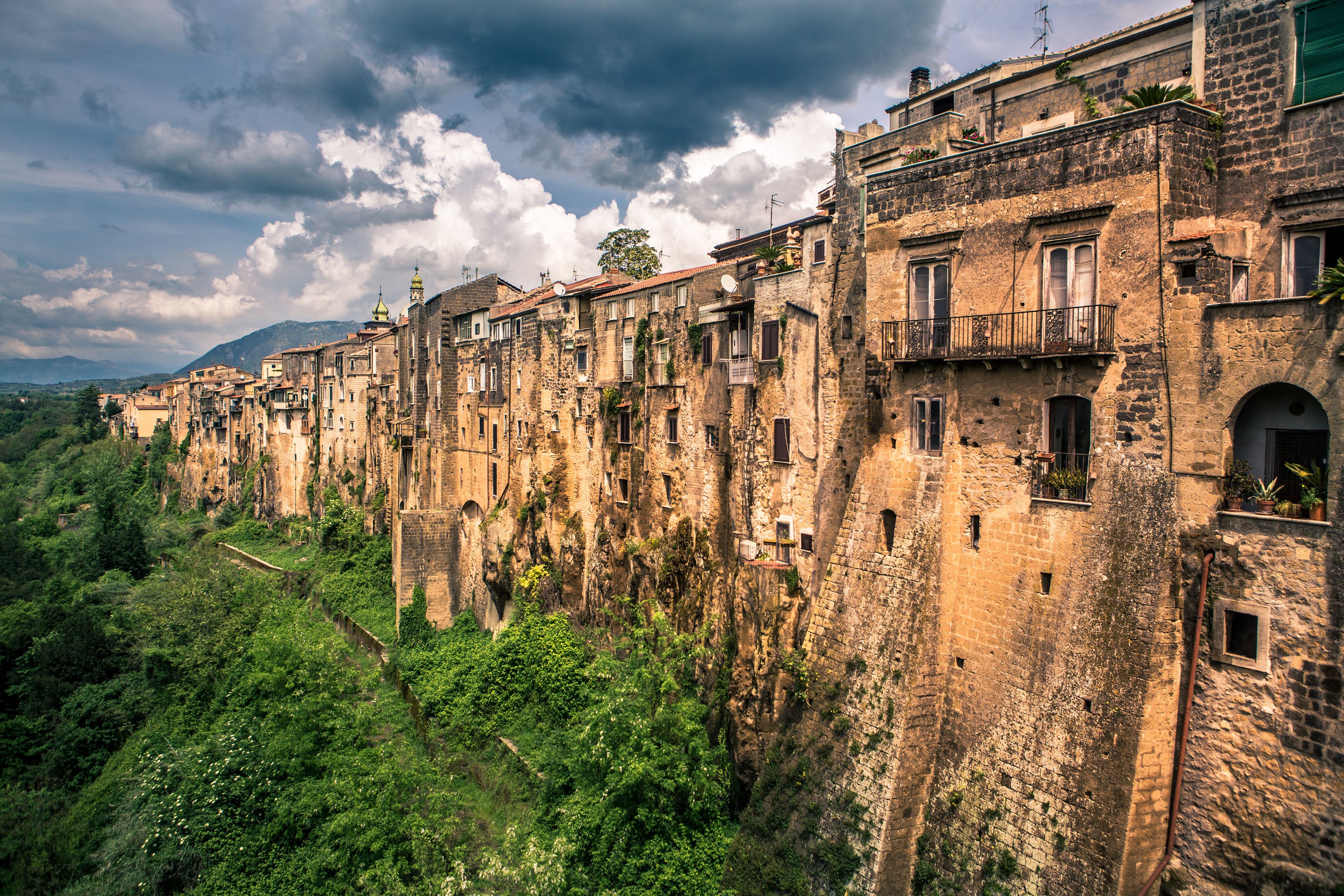 A row of historic stone buildings with balconies, set against a backdrop of lush greenery and a cloudy sky.