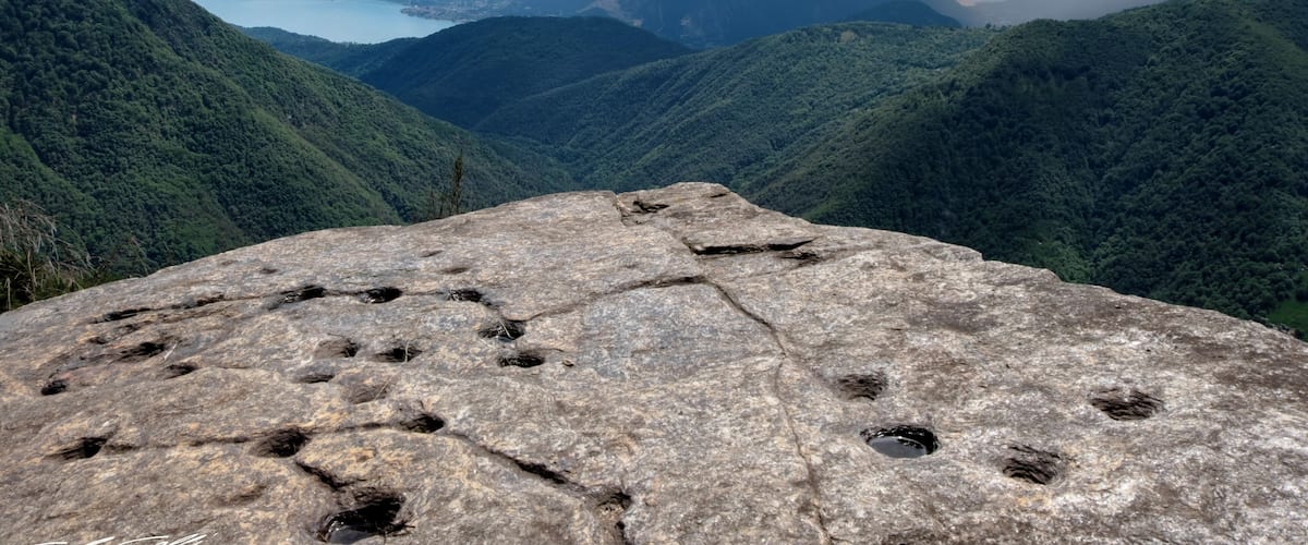 A rocky outcrop with multiple animal tracks, surrounded by lush green hills and a distant lake under a partly cloudy sky.