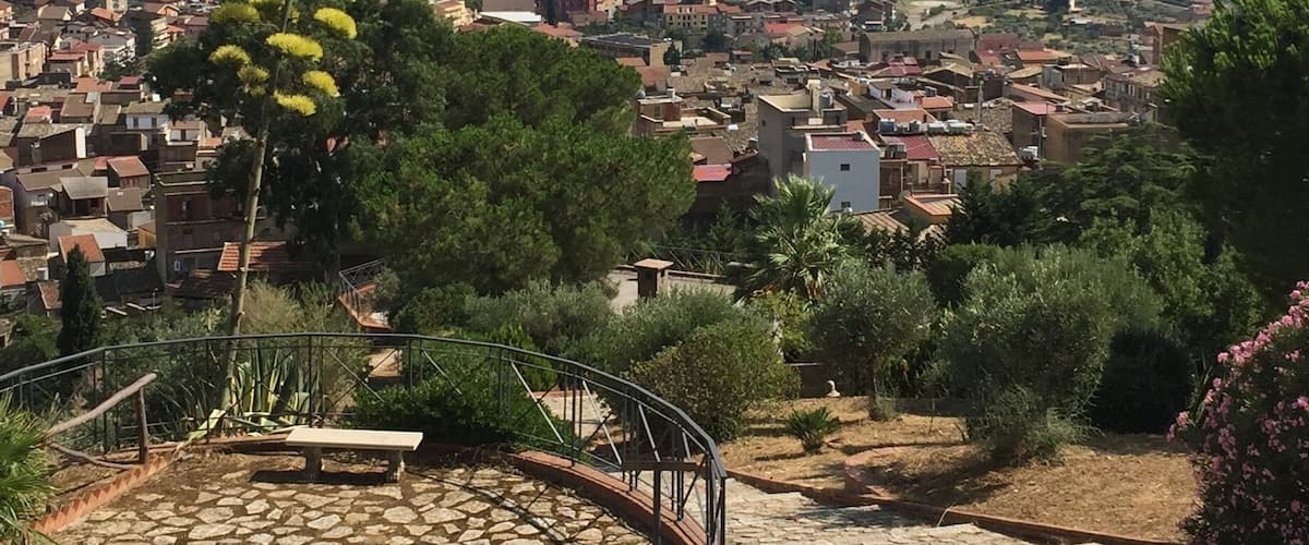 A cobblestone plaza with a bench, surrounded by greenery and a view of a town with buildings and mountains in the background.