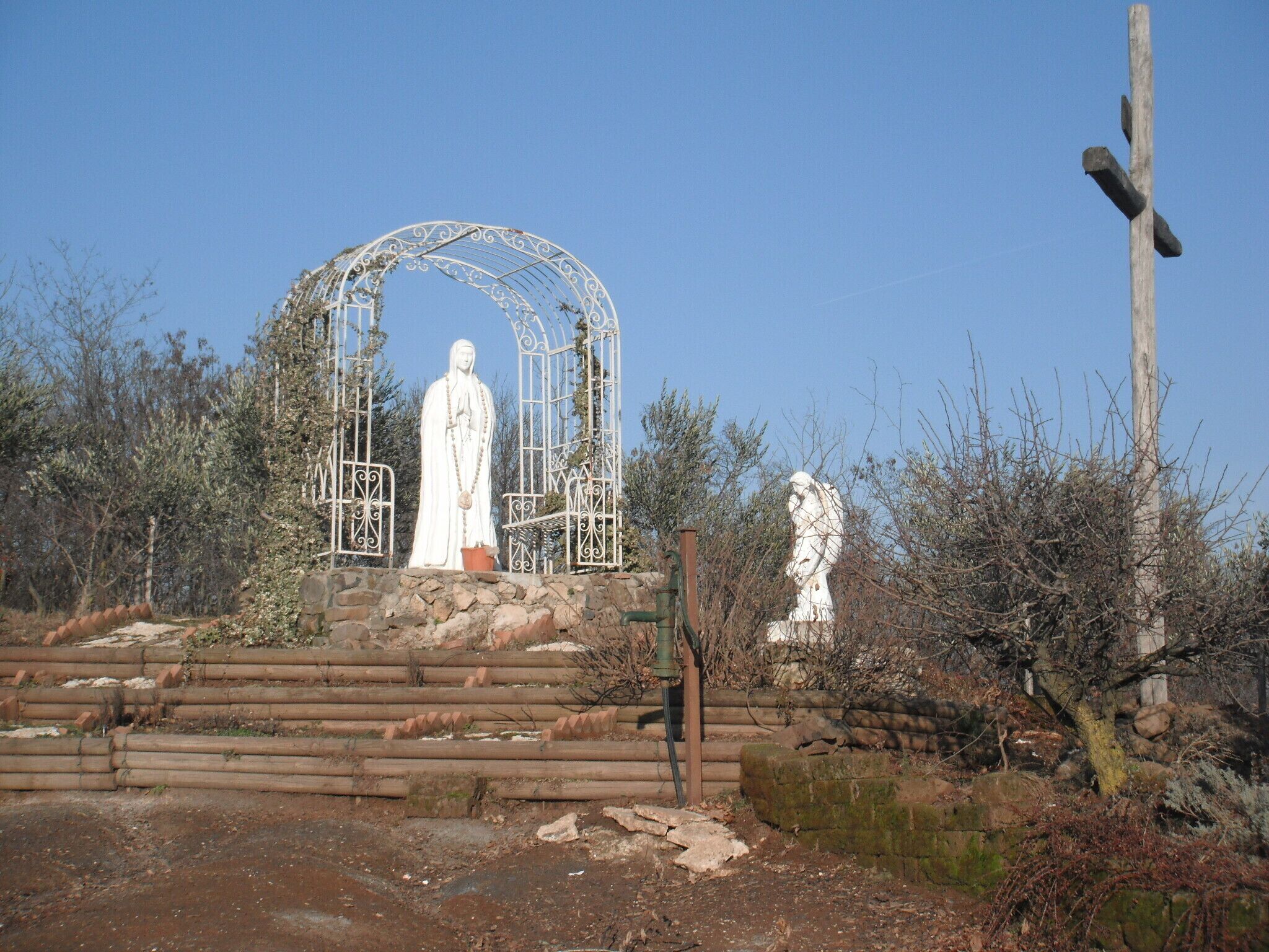 A white statue of a robed figure in a metal archway, surrounded by trees and a wooden fence.