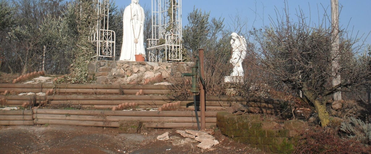 A white statue of a robed figure in a metal archway, surrounded by trees and a wooden fence.