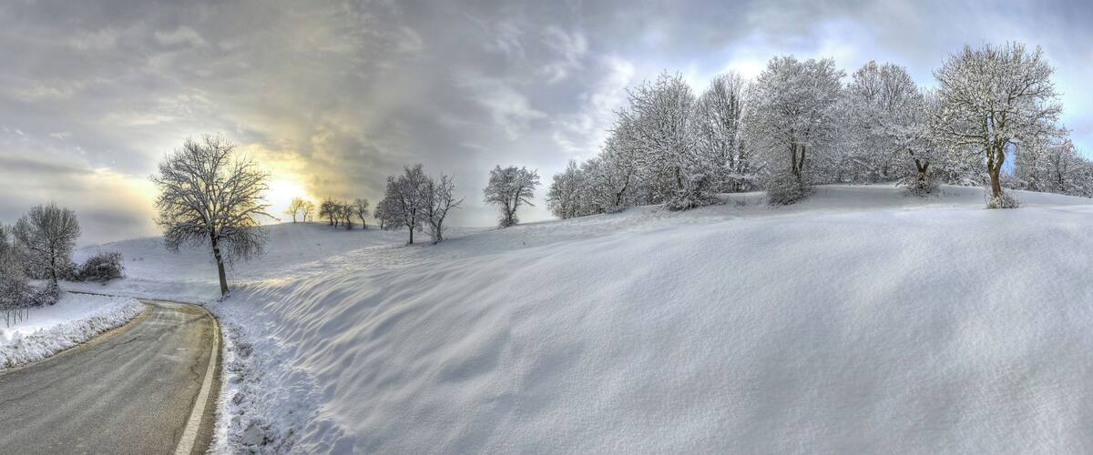 A snow-covered landscape with a winding road, trees, and a cloudy sky.