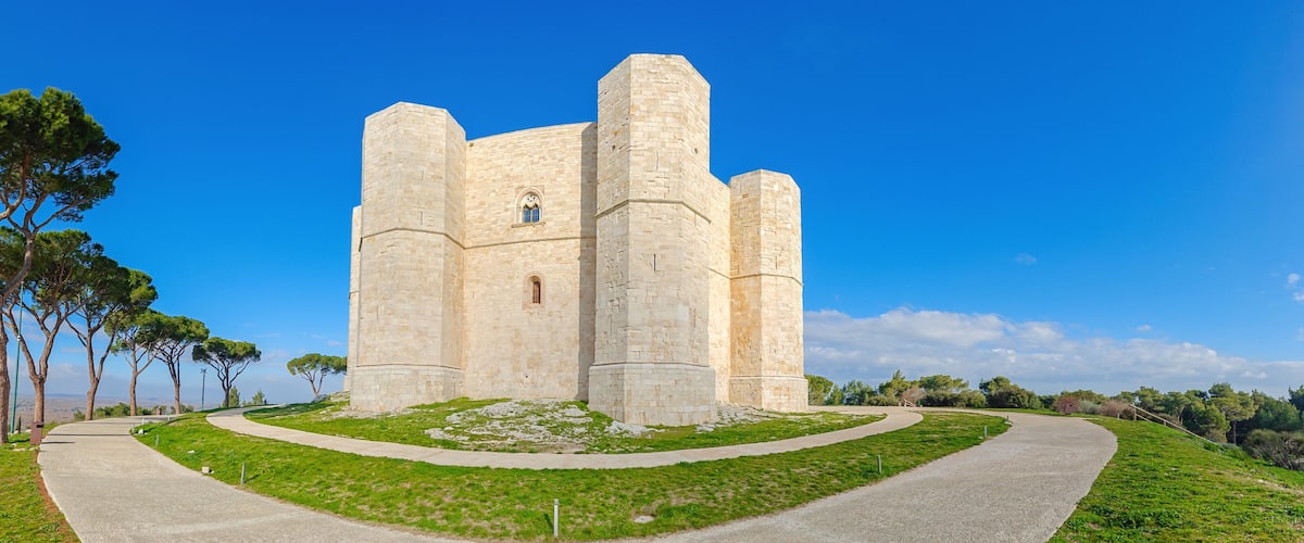 A historic stone castle with a circular tower, surrounded by a grassy area and tall trees.