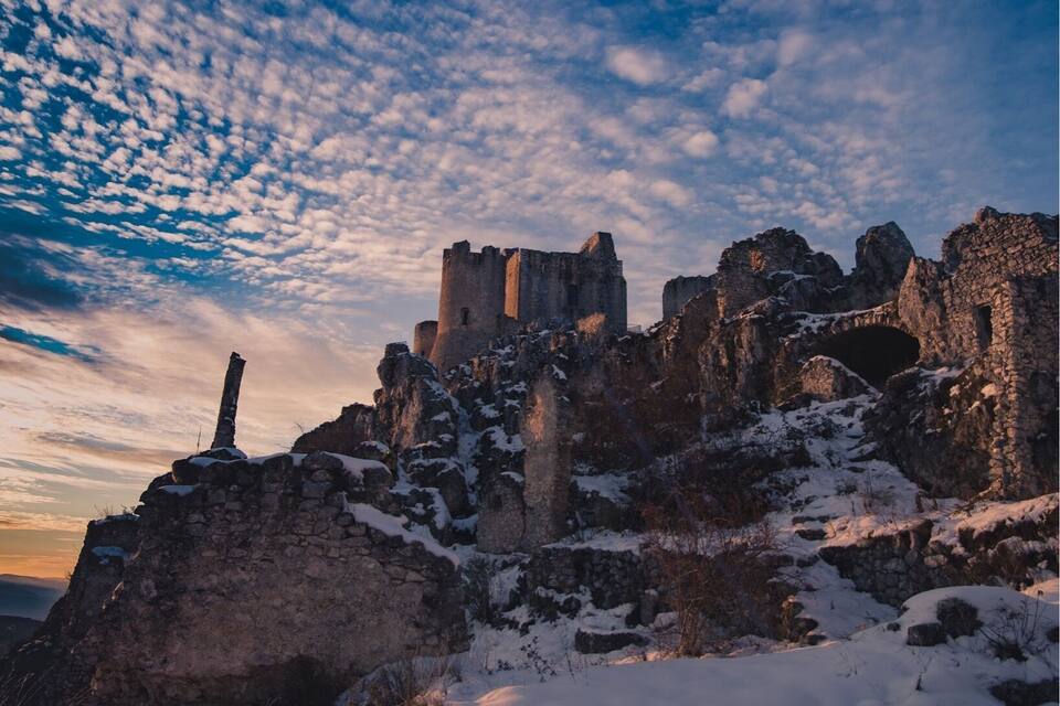 En snötäckt ruin med ett slott i bakgrunden under en molnig himmel.