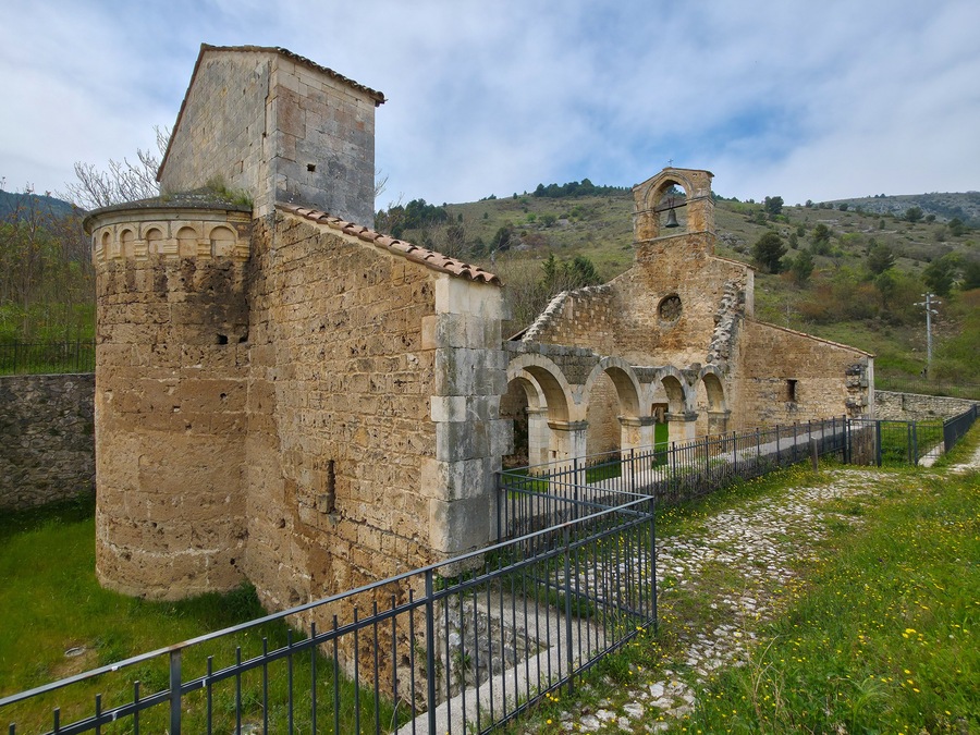 An ancient stone building with a bell tower, surrounded by a stone wall and a metal fence.