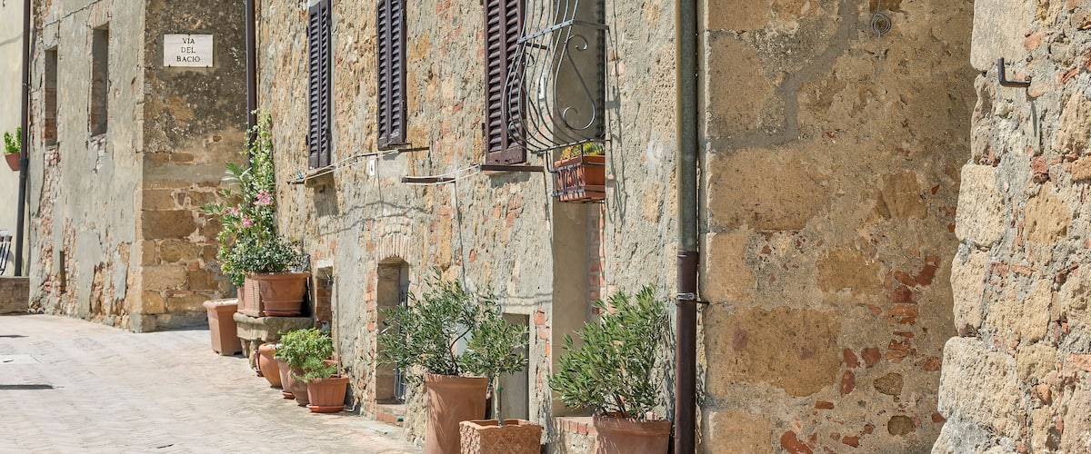 A narrow street with old stone buildings, potted plants, and signs indicating 'VIA DEL BACIO' and 'VIA BUIA'.