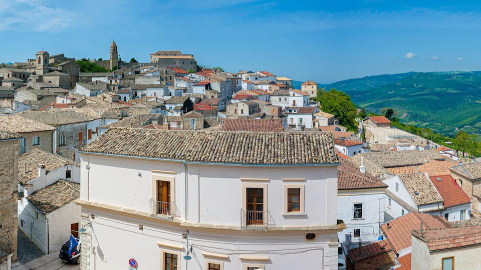A historic town with terracotta-tiled rooftops, a prominent bell tower, and rolling green hills in the background.
