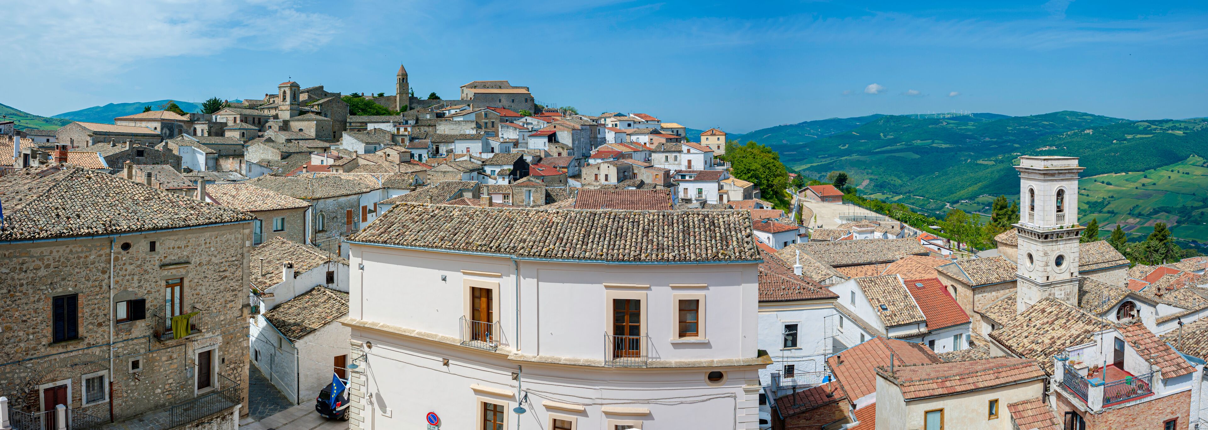 A historic town with terracotta-tiled rooftops, a prominent bell tower, and rolling green hills in the background.