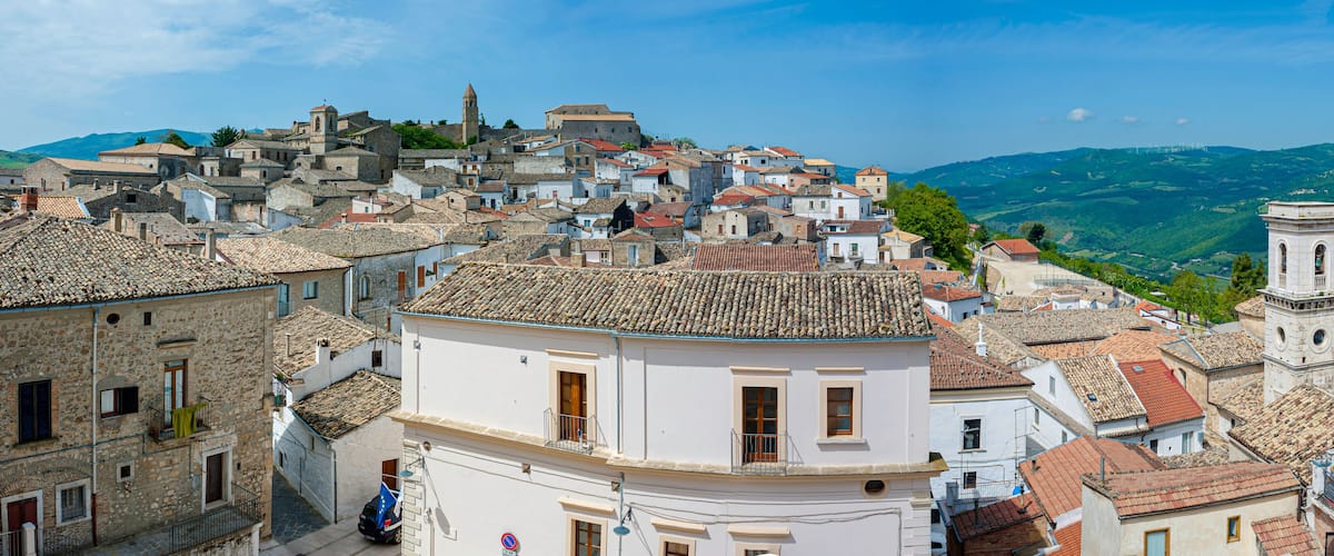 A historic town with terracotta-tiled rooftops, a prominent bell tower, and rolling green hills in the background.