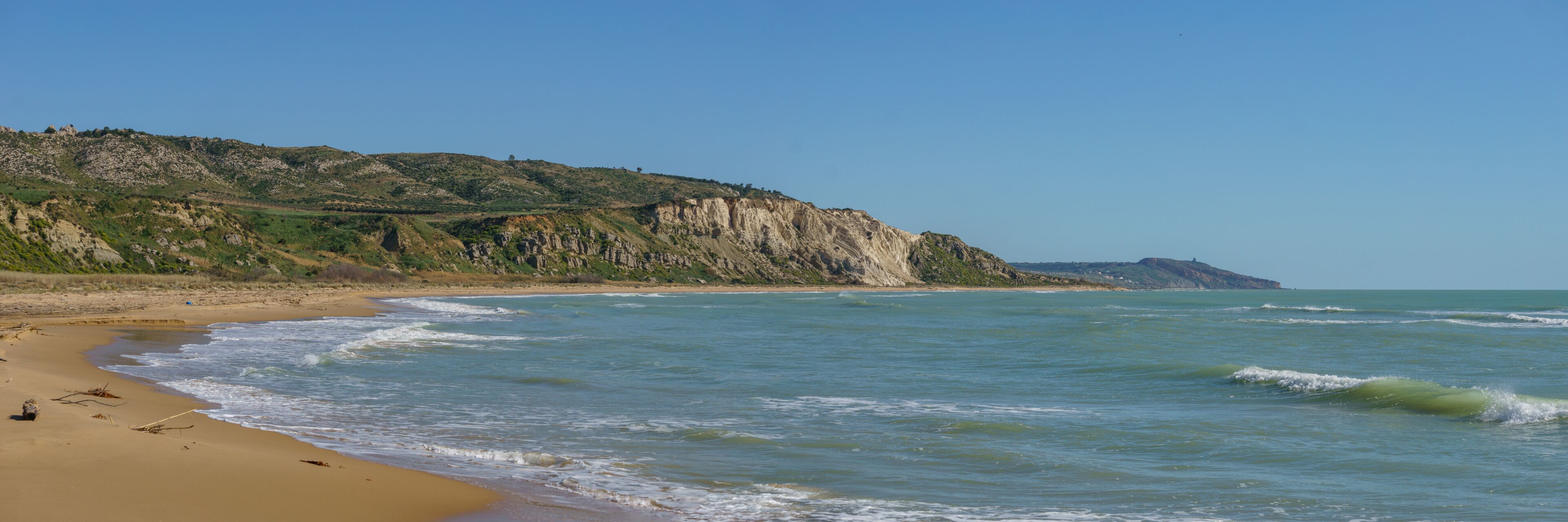A sandy beach with gentle waves, a rocky cliff, and a clear blue sky.