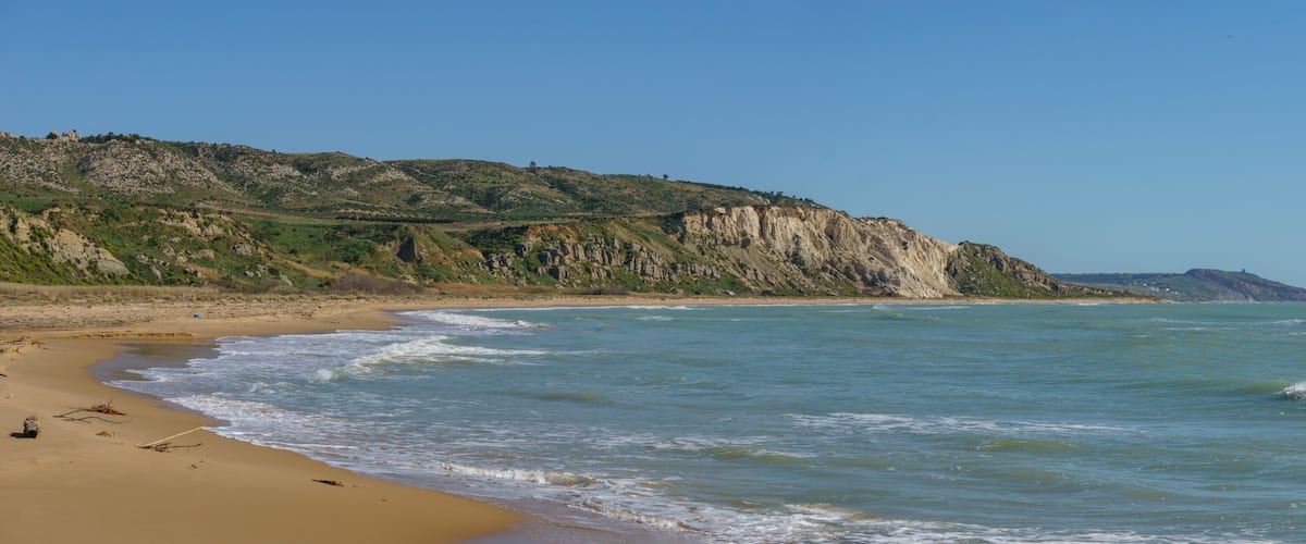 A sandy beach with gentle waves, a rocky cliff, and a clear blue sky.