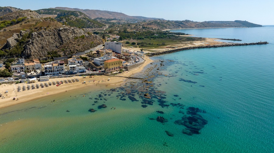 A coastal town with a beach, clear blue waters, rocky outcrops, and a hilly backdrop.