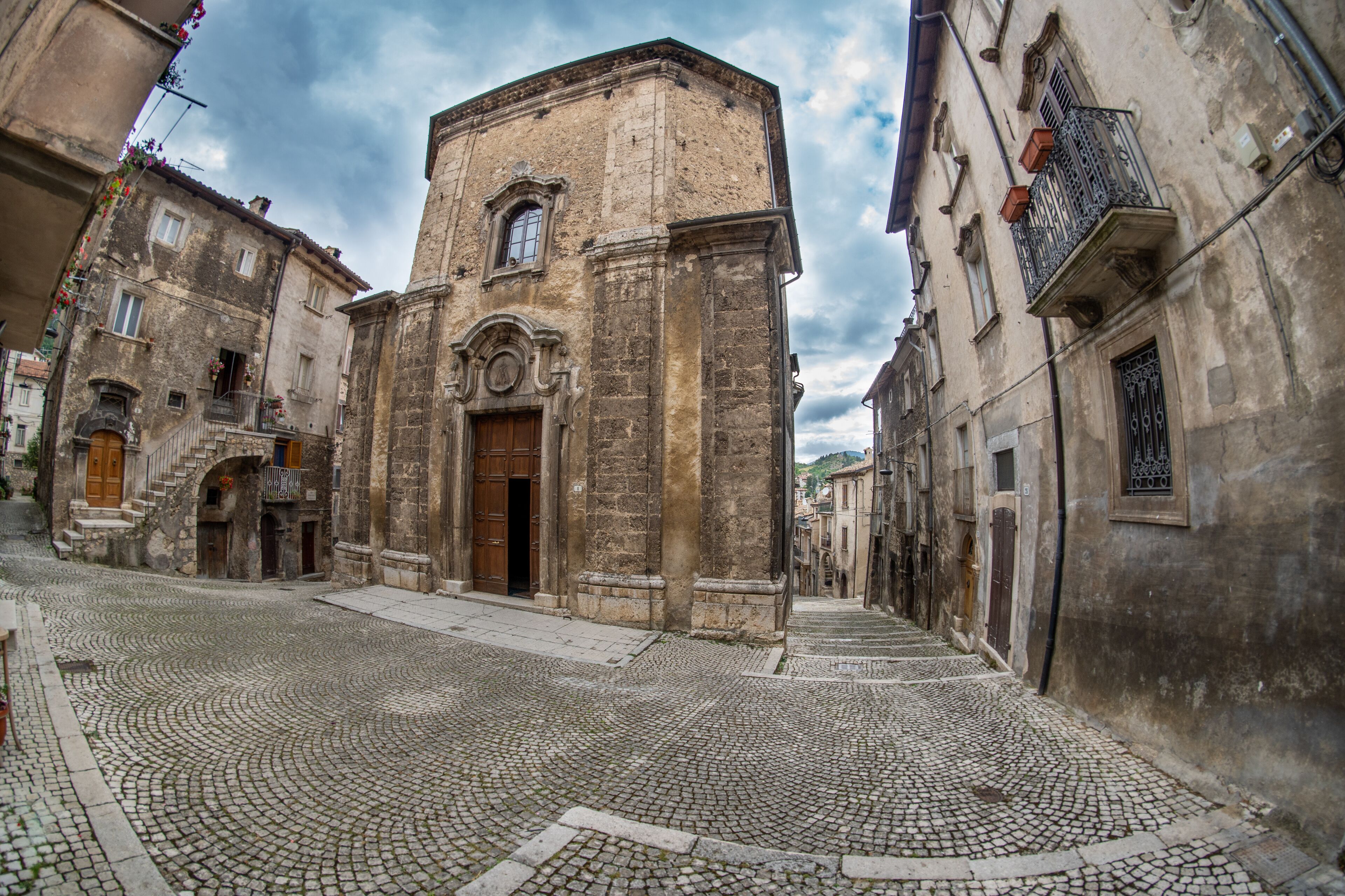A cobblestone street flanked by historic buildings with a prominent church featuring a wooden door and a bell tower.