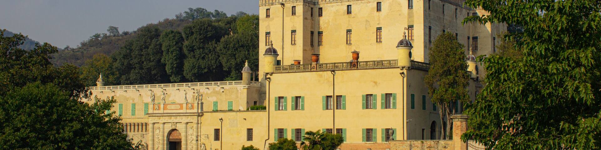 A large, historic building with green shutters, surrounded by lush greenery and hills in the background.