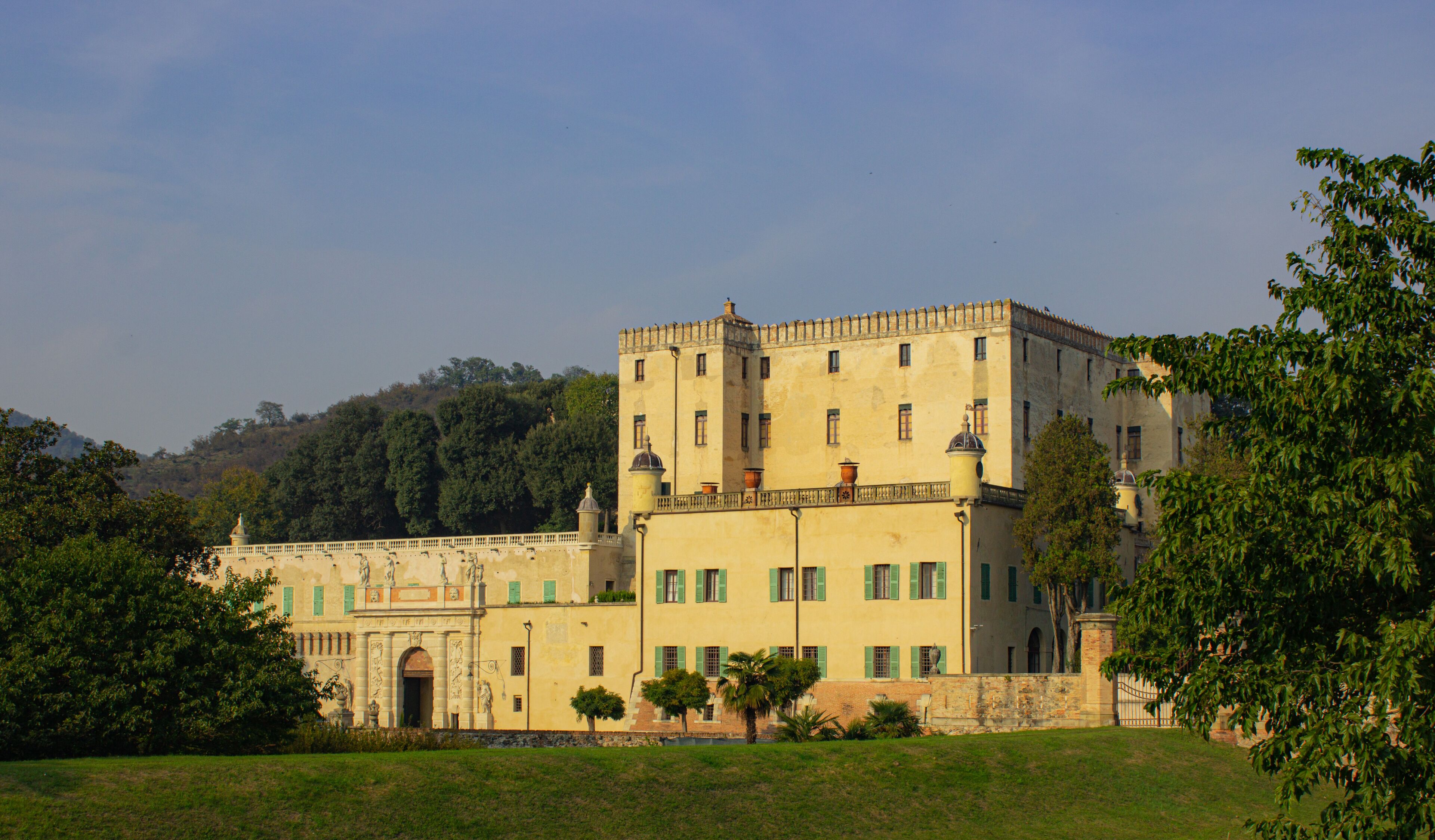 A large, historic building with green shutters, surrounded by lush greenery and hills in the background.
