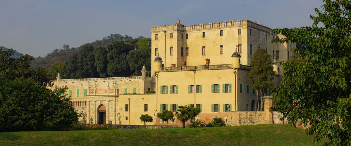 A large, historic building with green shutters, surrounded by lush greenery and hills in the background.
