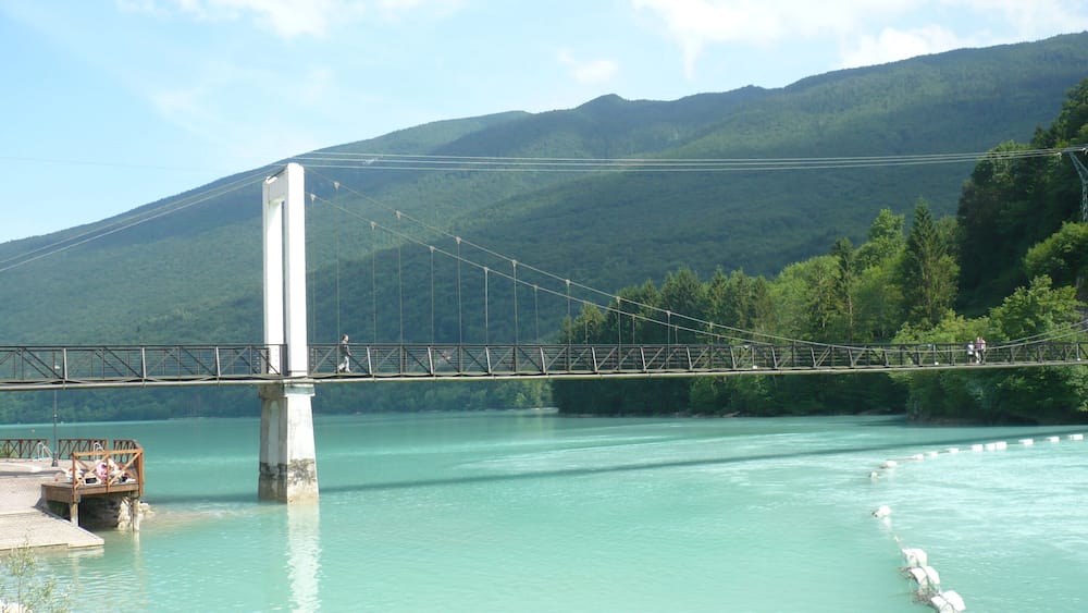 Eine Hängebrücke über türkisfarbenem Wasser, im Hintergrund Berge.