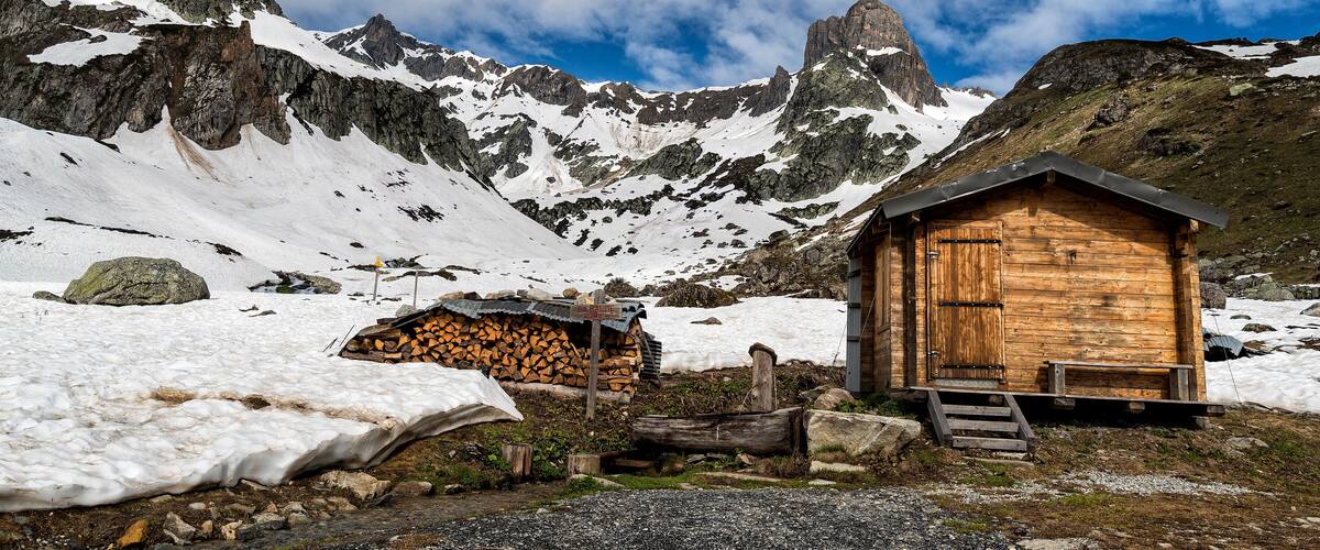 A wooden mountain hut with a snow-covered landscape and rocky peaks in the background.