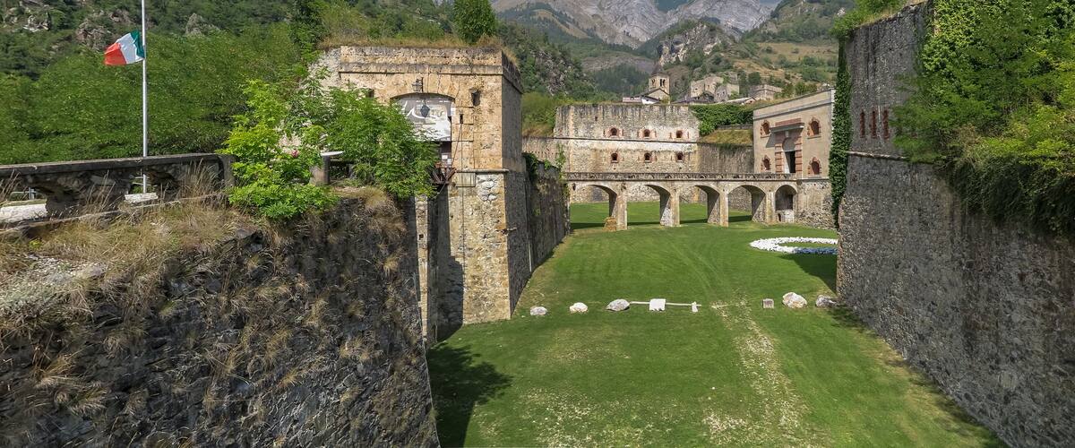 A historic stone fortress with a flag, lush greenery, and mountains in the background.