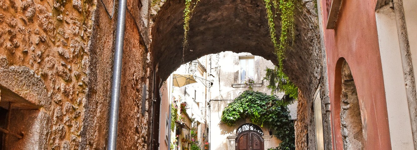 A cobblestone alleyway with an archway, stone walls, and a staircase leading to a building with a wooden door.