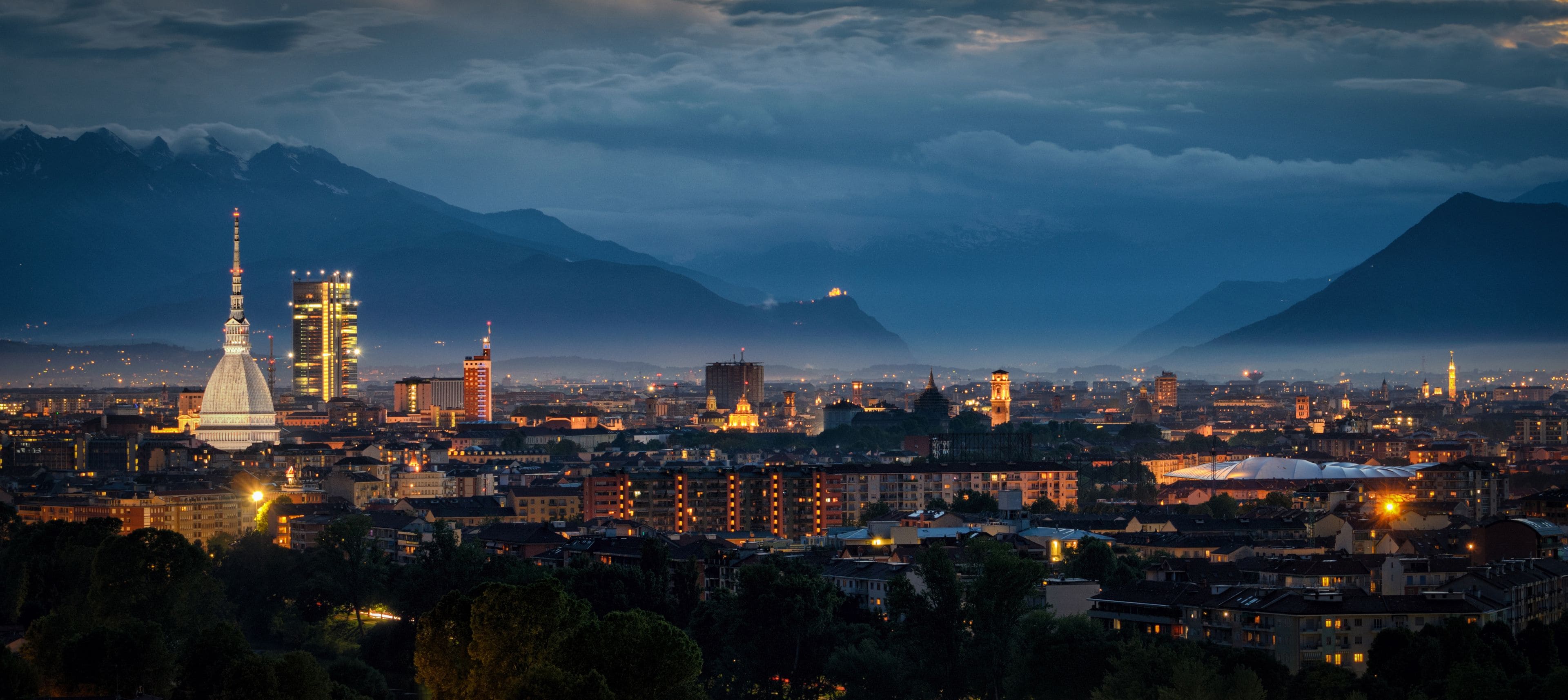 A cityscape at dusk with illuminated buildings and mountains in the background.