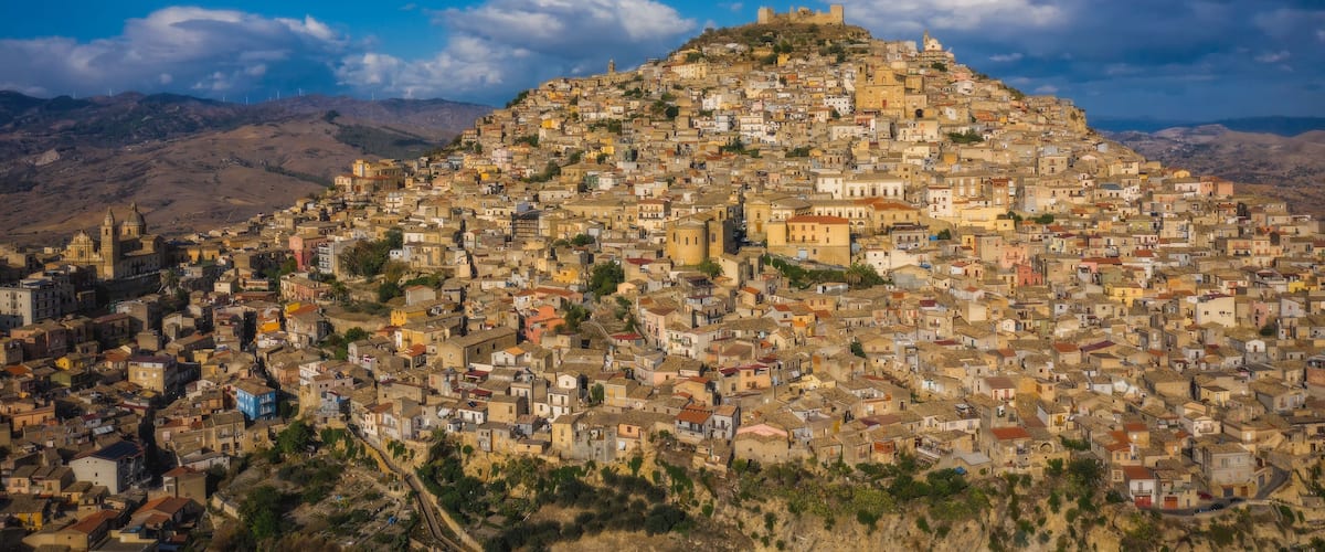 Un pueblo pintoresco y concurrido asentado en una ladera, con arquitectura histórica y un castillo imponente en la cima, bajo un cielo azul despejado.