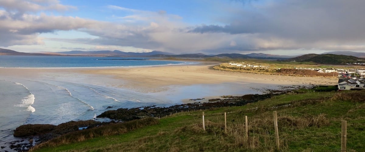 A coastal landscape with a sandy beach, rocky shoreline, grassy hills, and a small settlement with houses.