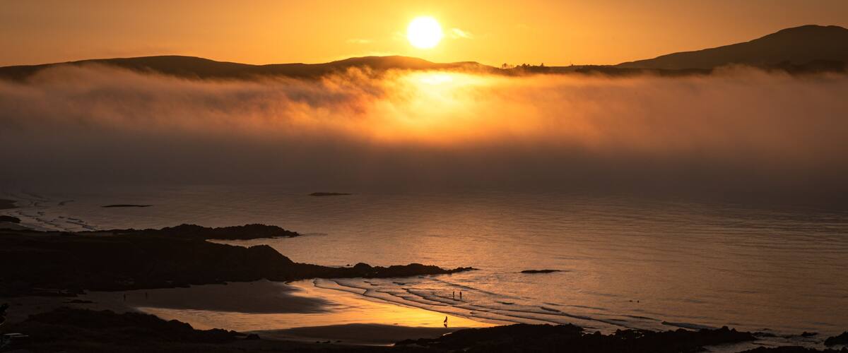 A sunset over a coastal landscape with a beach, rocky outcrops, and a mountain in the background.