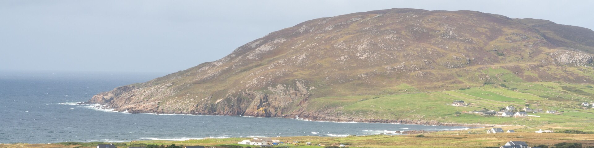 A coastal village with houses on rolling hills and a large mountain in the background.