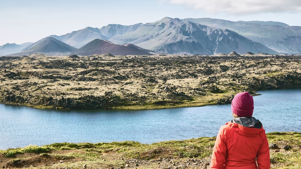 A person in a red jacket and pink beanie overlooking a lake with mountains in the background.