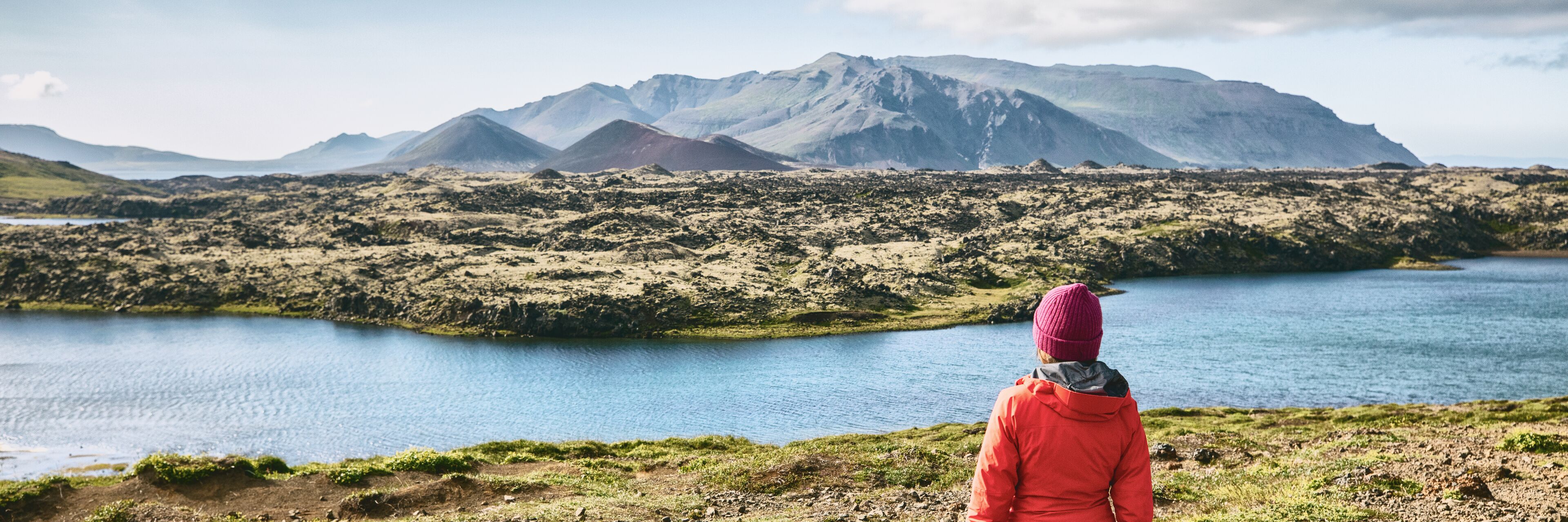 A person in a red jacket and pink beanie overlooking a lake with mountains in the background.