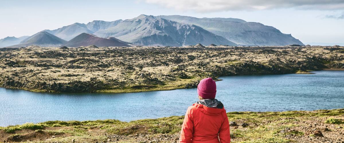 A person in a red jacket and pink beanie overlooking a lake with mountains in the background.