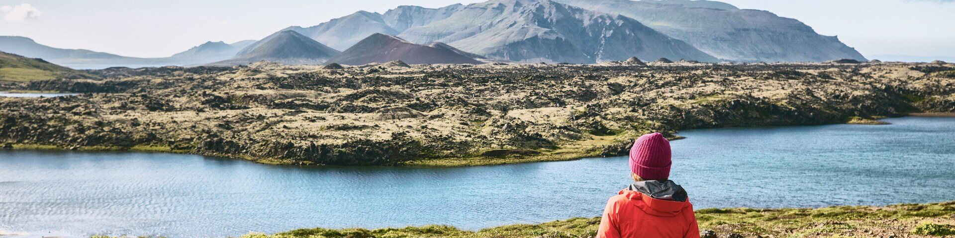 A person in a red jacket and pink beanie overlooking a lake with mountains in the background.