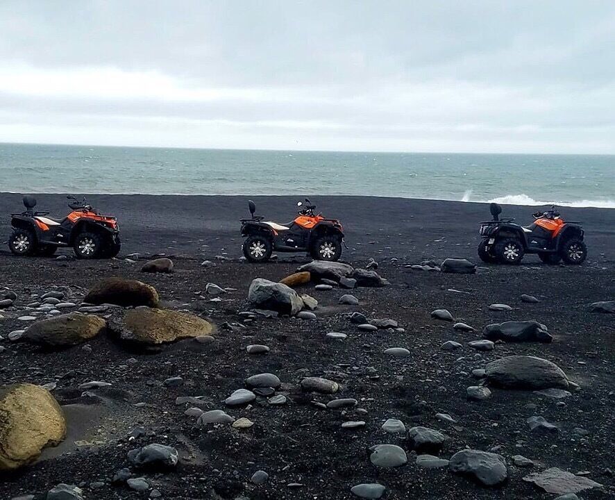 Three orange ATVs parked on a rocky beach with the ocean in the background.