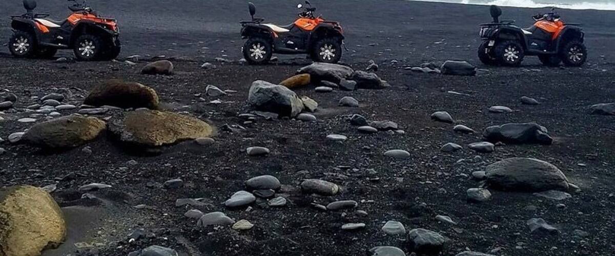 Three orange ATVs parked on a rocky beach with the ocean in the background.