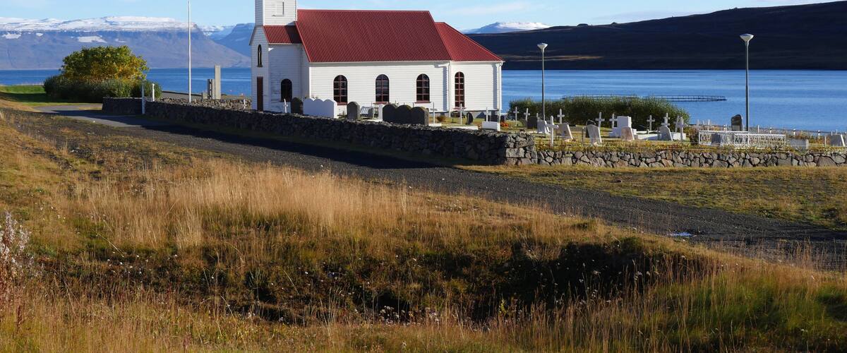 A white church with a red roof by a lake, surrounded by graves and a stone wall.