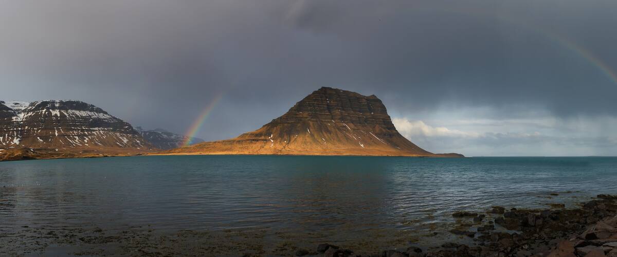 A coastal landscape with a prominent rock formation, a calm sea, and two rainbows.