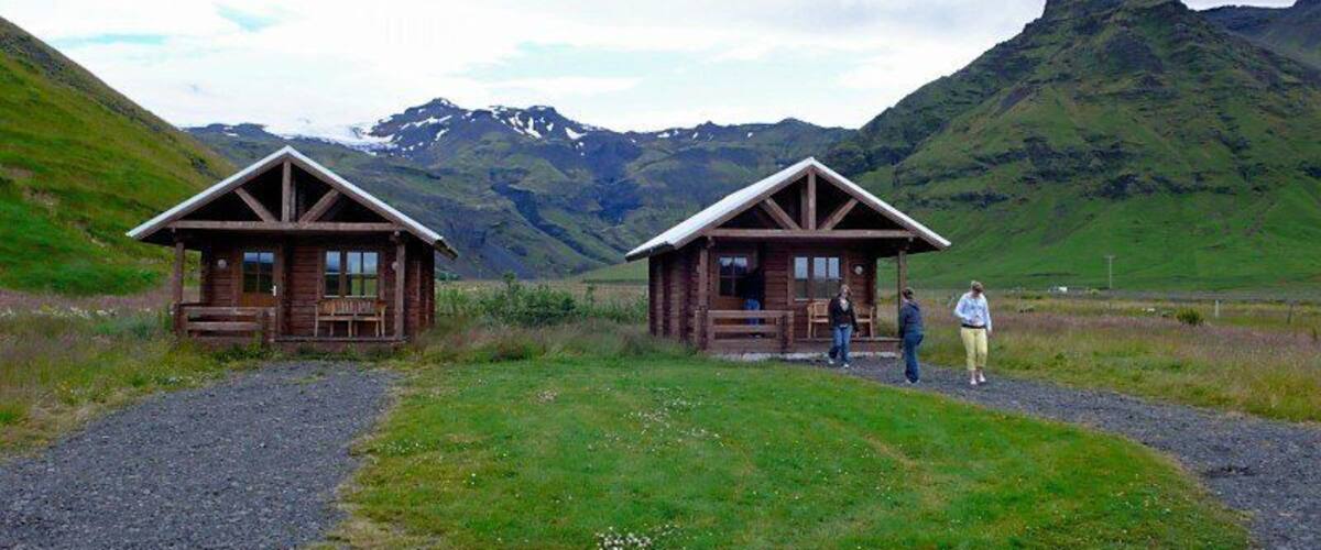 Two wooden cabins with a gravel path leading to them, set against a mountainous backdrop.
