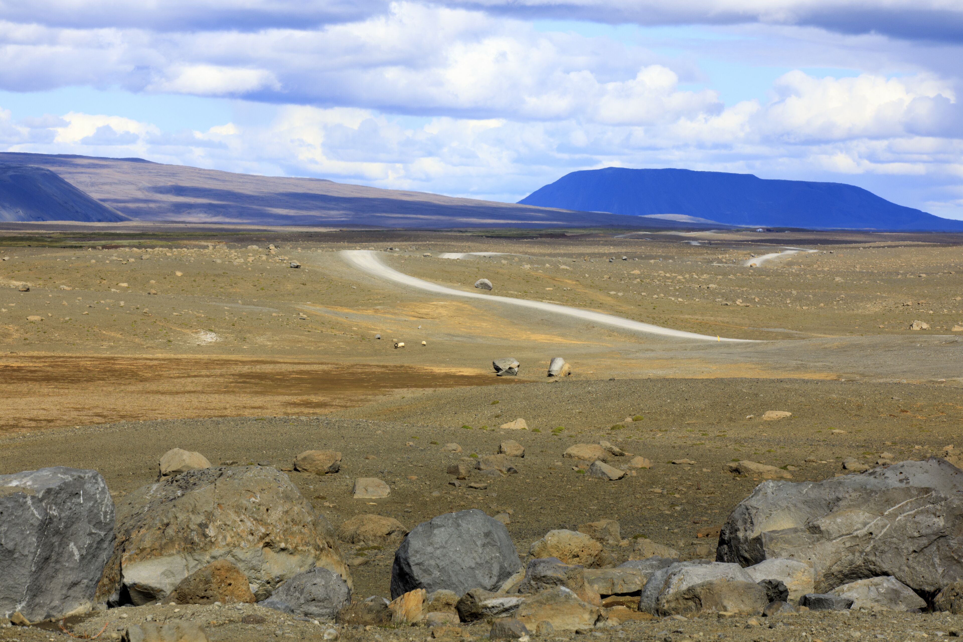 A barren landscape with a winding road, large rocks, and distant mountains under a cloudy sky.