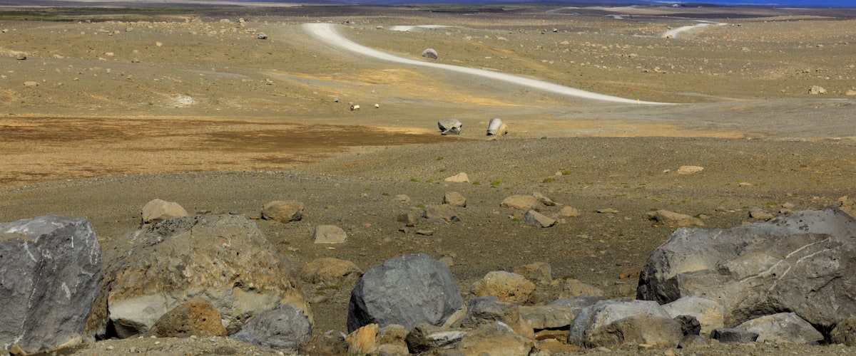 A barren landscape with a winding road, large rocks, and distant mountains under a cloudy sky.