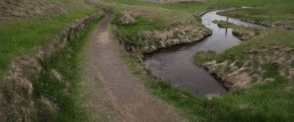 A narrow dirt path beside a meandering stream in a grassy landscape with rolling hills in the background.
