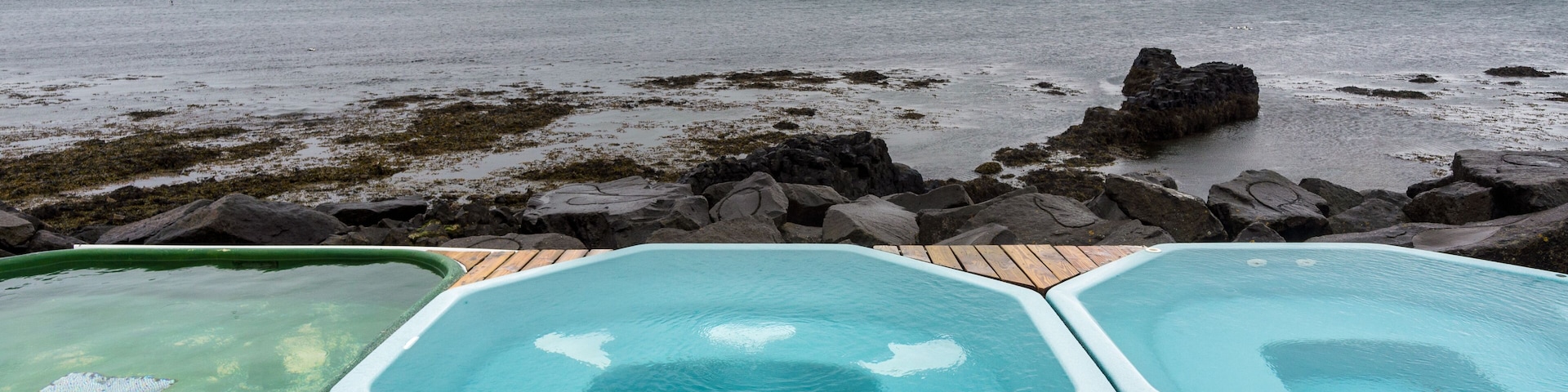 Una terraza de madera con tres tinas de hidromasaje con vista a una costa rocosa y al mar.